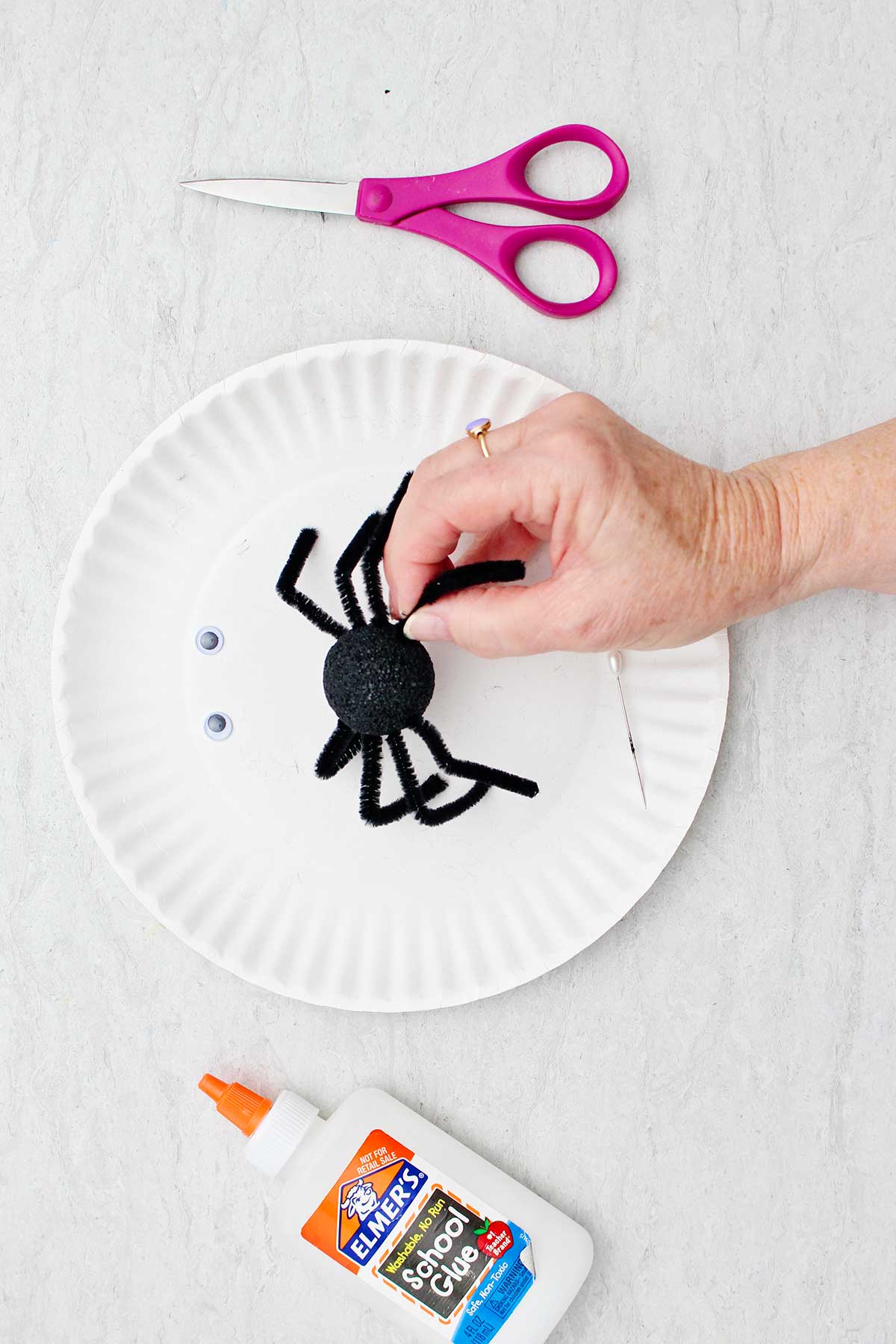 Hand attaching black pipe cleaner legs to a black styrofoam ball to make spider with other supplies near by.