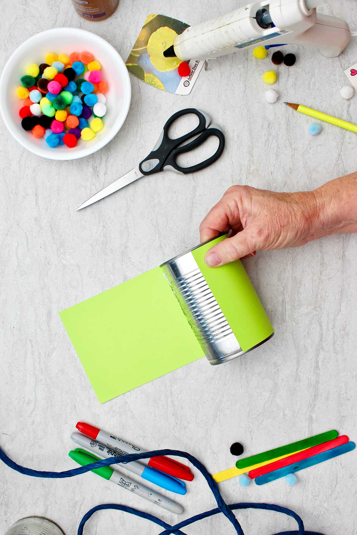 Person securing a strip of green paper around an aluminum can with other supplies for decoration near by.