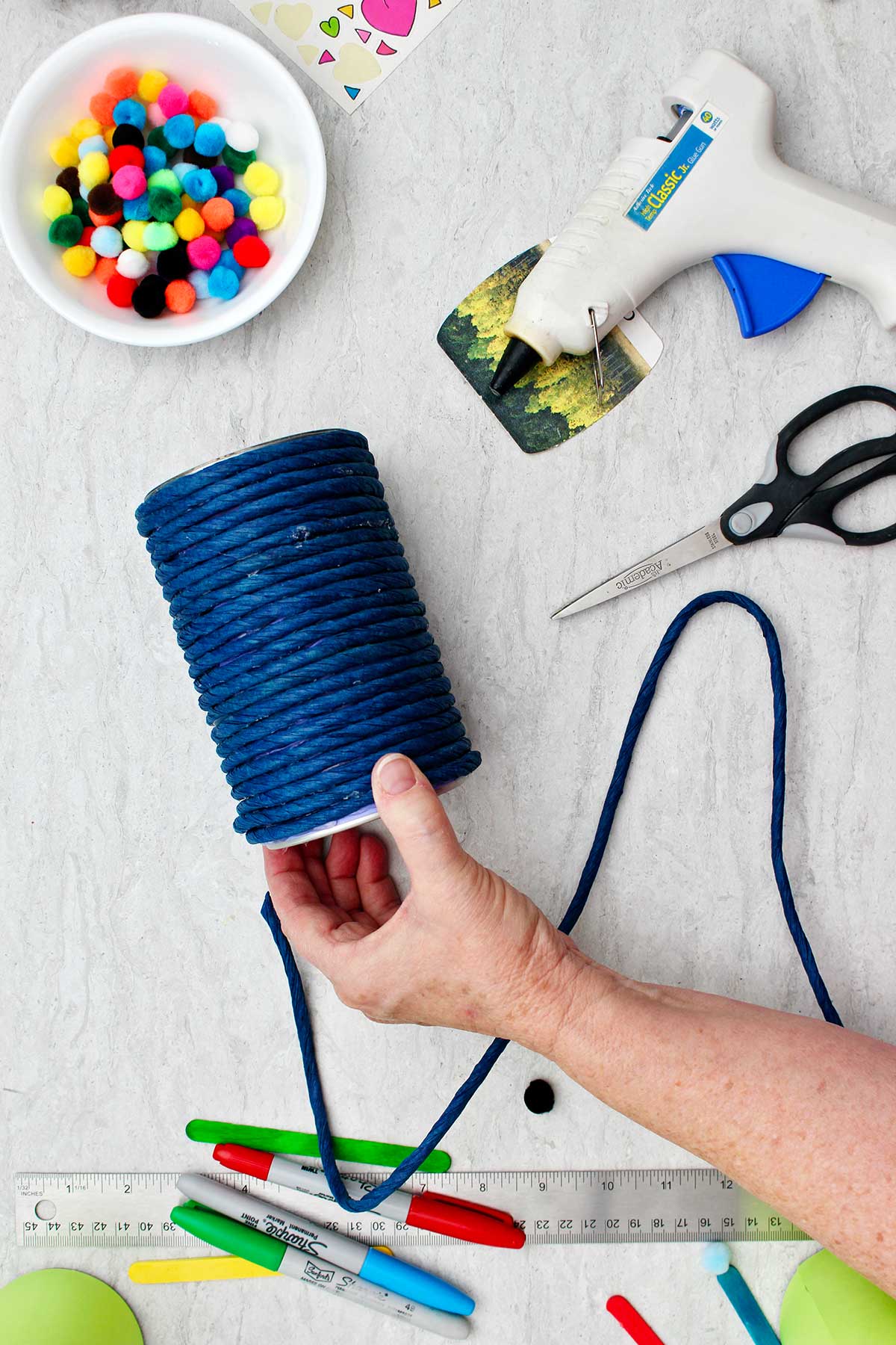 Person hot gluing thick blue twine around a can to make a pencil and pen holder with other supplies for decoration near by.