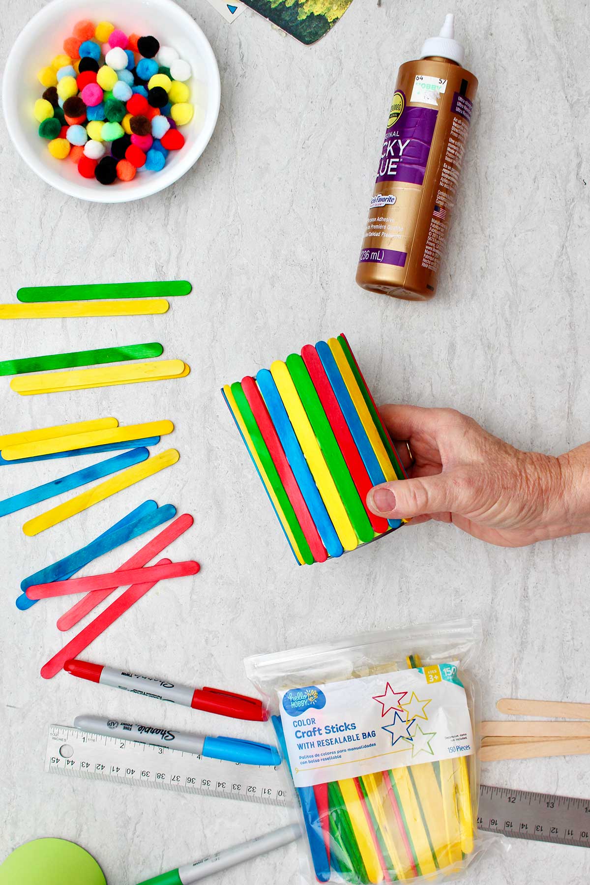 Person holding completed pen and pencil holder decorated with colorful popsicle sticks with other supplies for decoration near by.