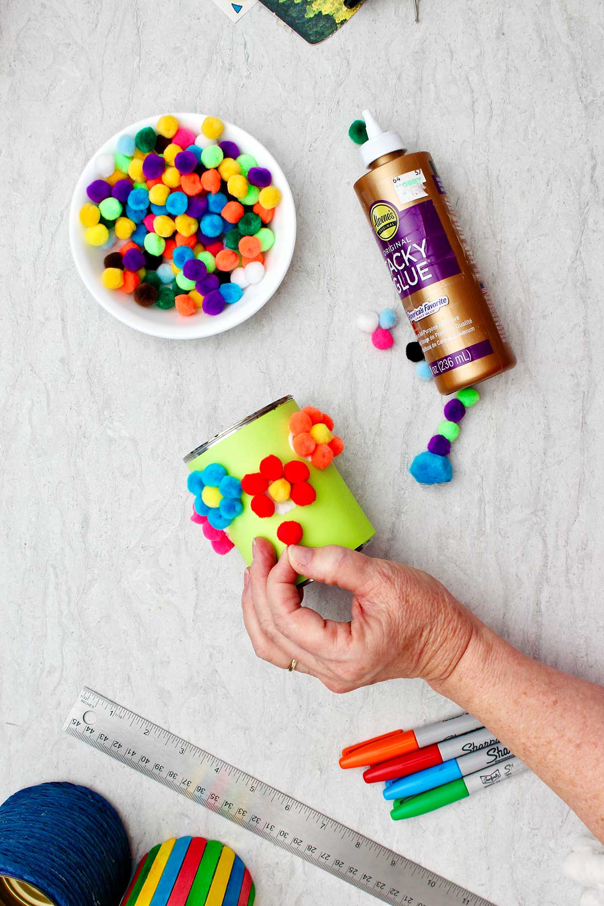 Person gluing a red pom pom to a paper covered can to make a flower with other supplies for decoration near by.