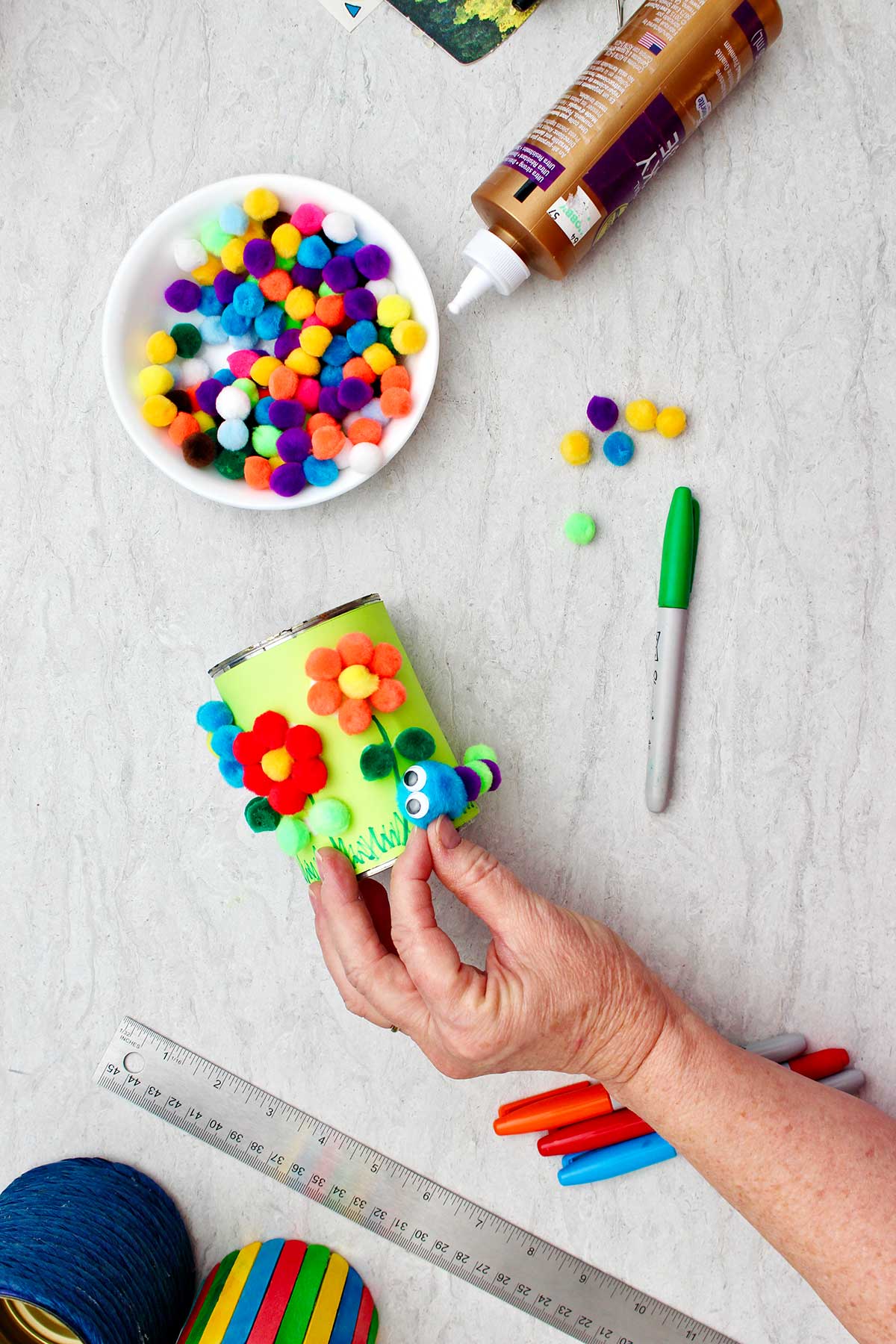 An image of a person securing a pompom to the pencil and pen holder to be the head of a caterpillar.