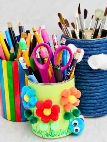 Three completed pencil and pen holders made from cans wrapped in blue twine, colorful paper and pom poms and multi colored popsicle sticks.