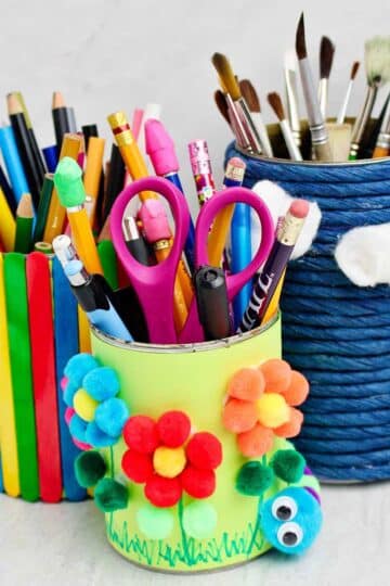 Three completed pencil and pen holders made from cans wrapped in blue twine, colorful paper and pom poms and multi colored popsicle sticks.