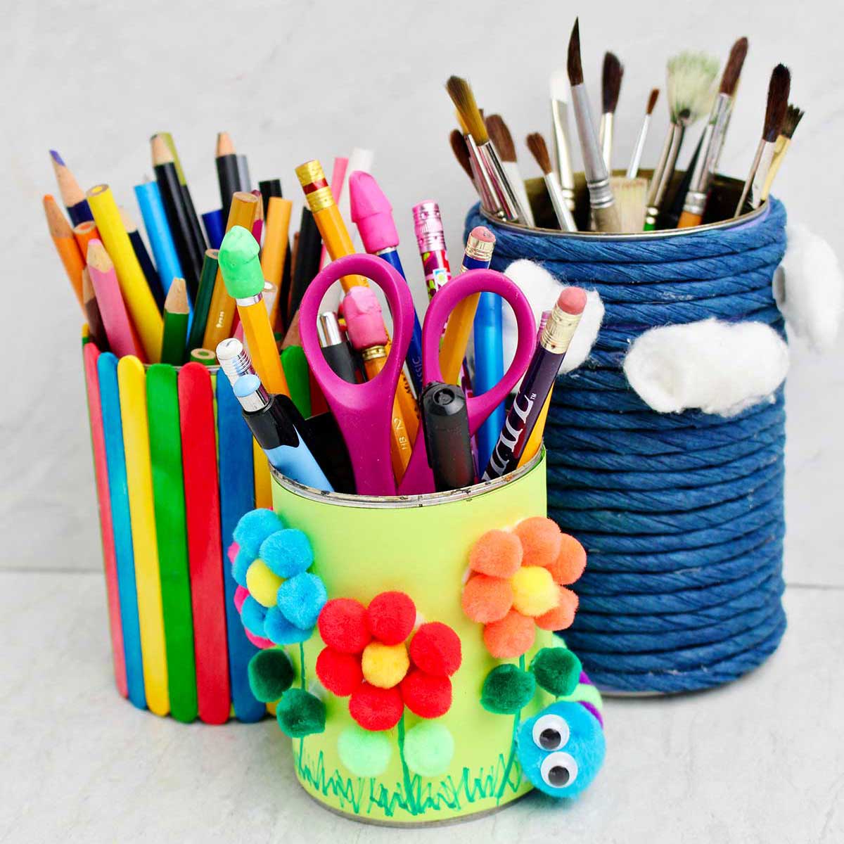 Three completed pencil and pen holders made from cans wrapped in blue twine, colorful paper and pom poms and multi colored popsicle sticks.