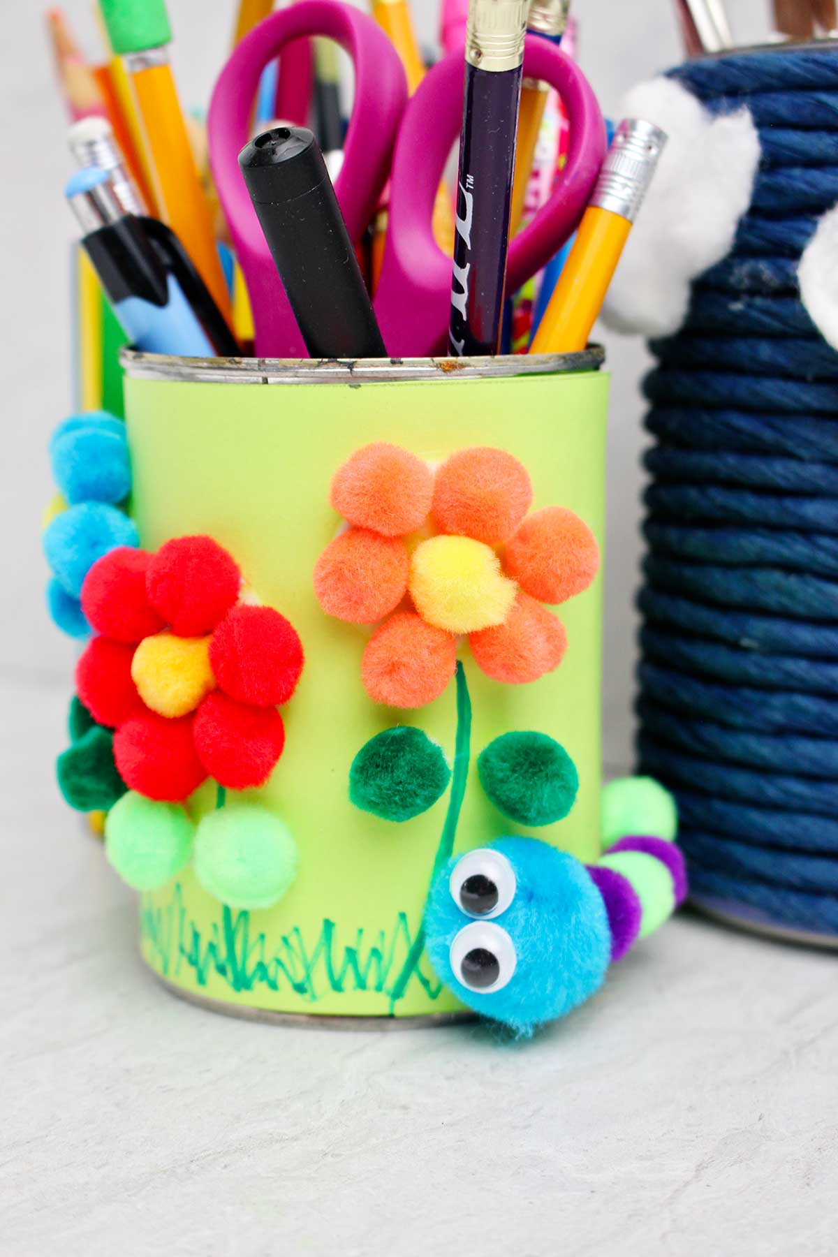 Close up of pen and pencil holder decorated with lime green paper and pom poms to make flowers and a caterpillar.