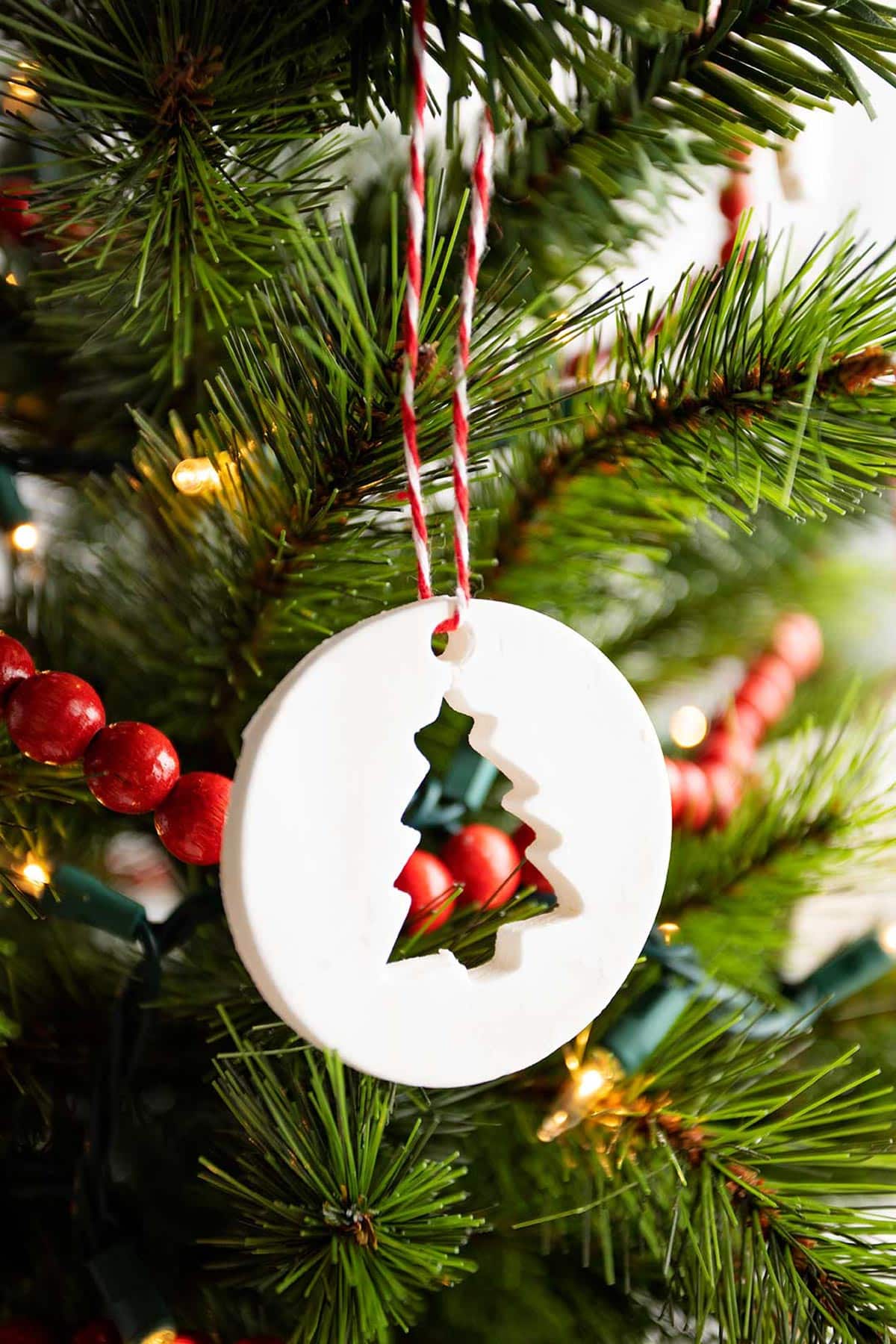 Circular ornament with tree cut-out hanging on a Christmas tree with white lights and red wooden garland.