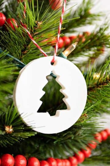 Person hanging circular cornstarch ornament with tree cut-out on a Christmas tree with white lights and red wooden garland.