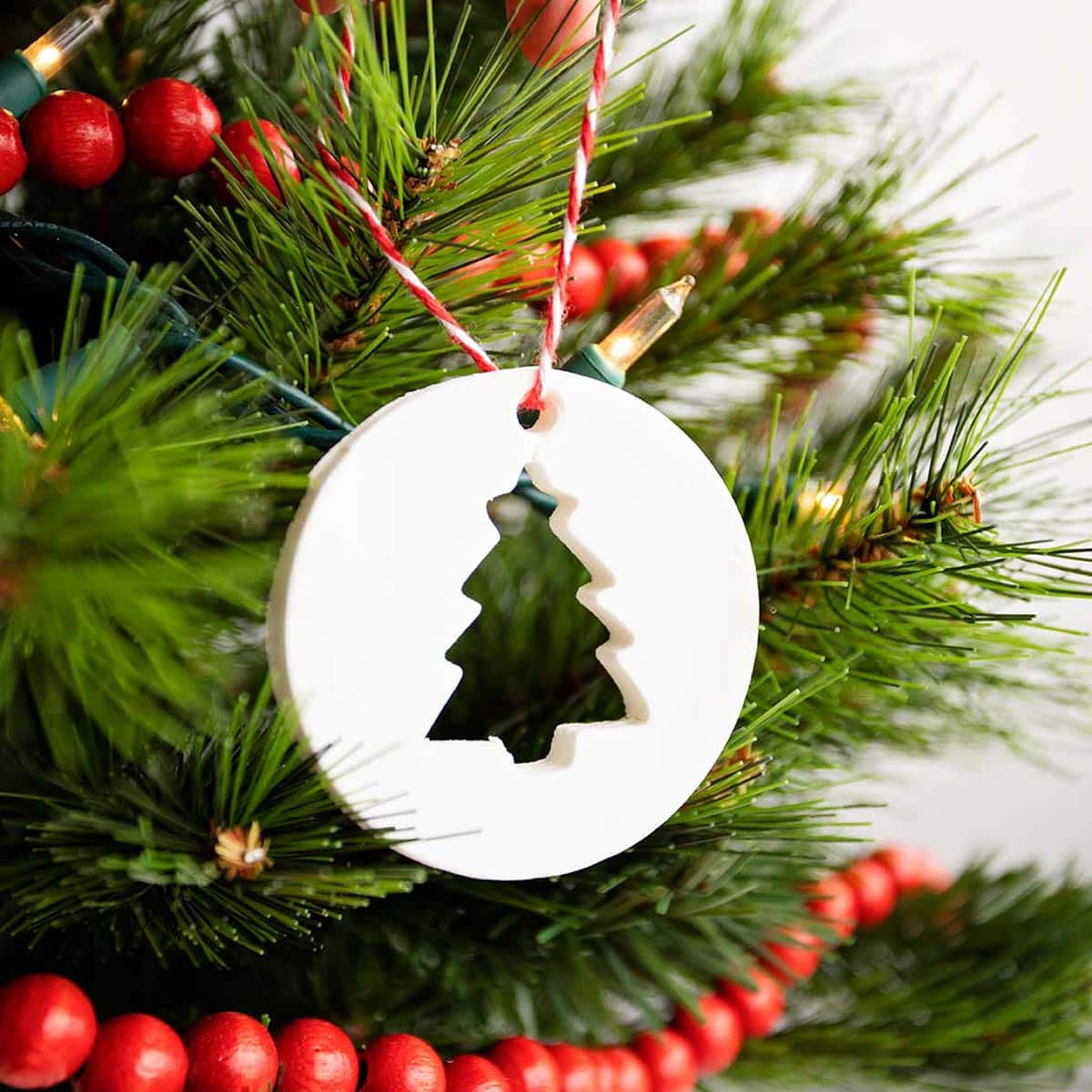 Person hanging circular cornstarch ornament with tree cut-out on a Christmas tree with white lights and red wooden garland.