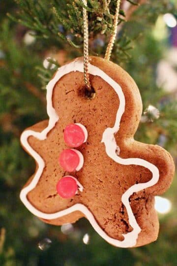 A gingerbread ornament decorated with red hots and white icing, hanging on the branch of a Christmas tree.