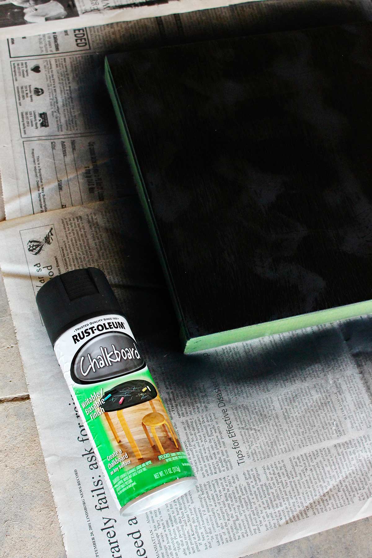 A spray painted piece of wood resting on newsprint with chalkboard spray paint near by.