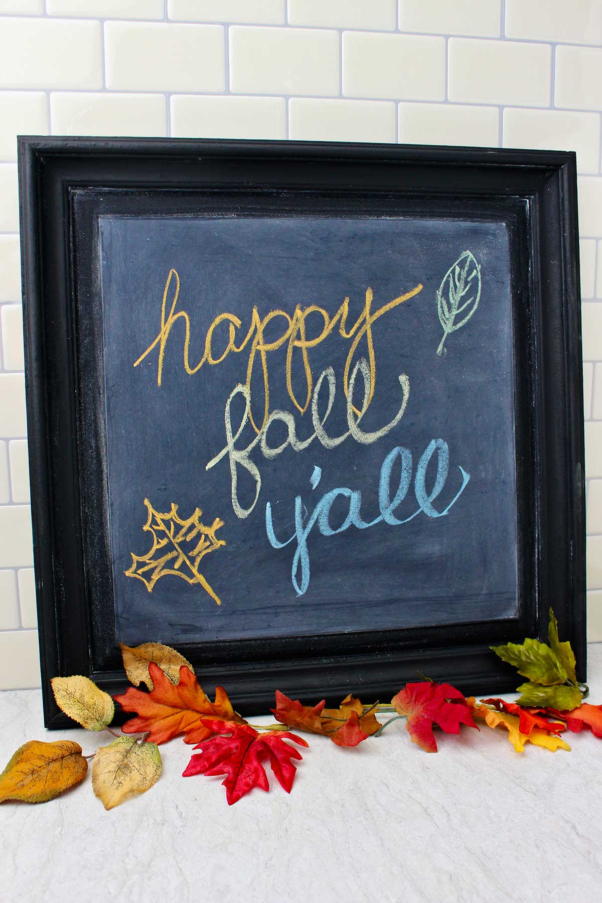 Black framed DIY chalkboard with "happy fall y'all" resting on a counter with fall leaves near by as decoration.