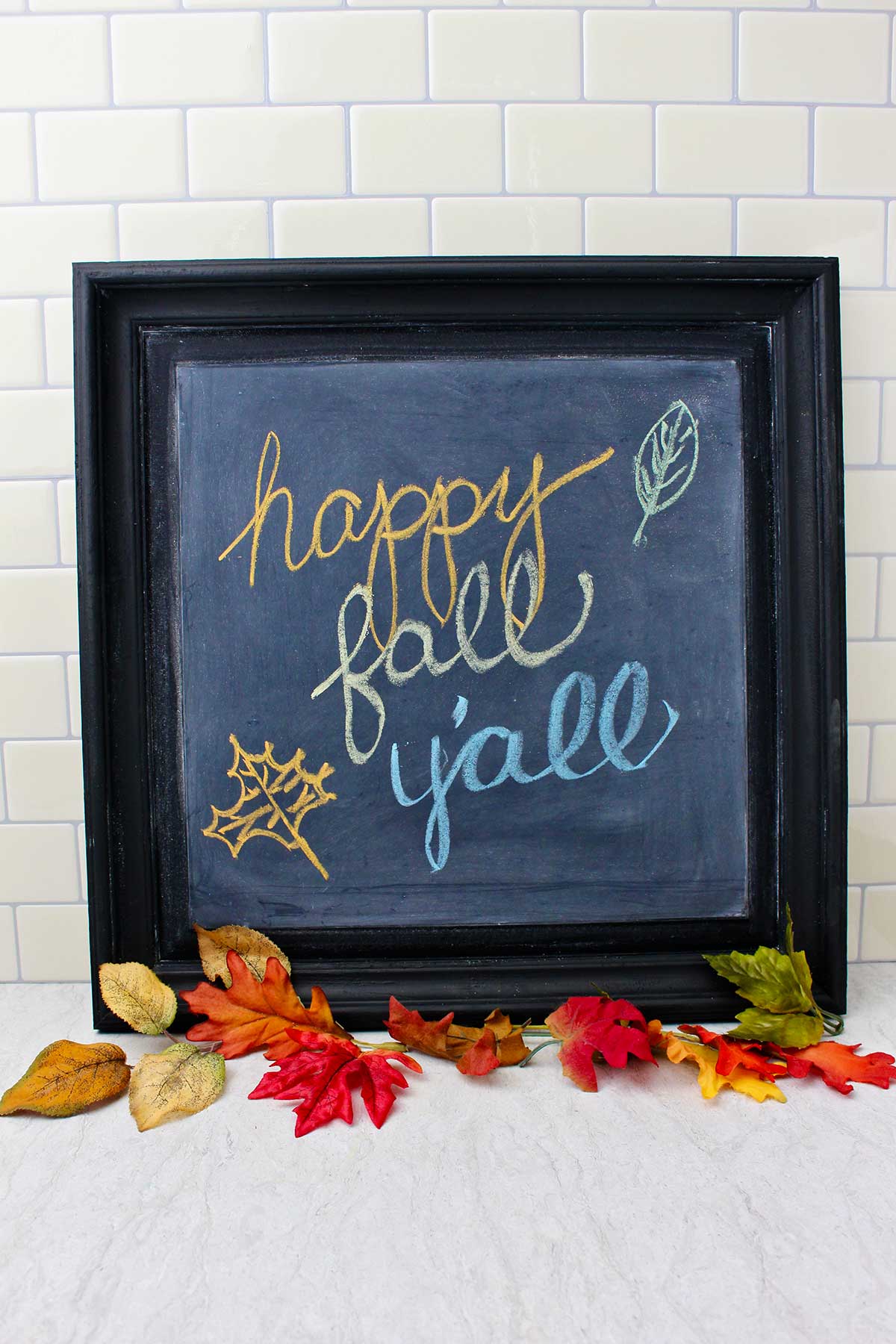 Black framed DIY chalkboard with "happy fall y'all" resting on a counter with fall leaves near by as decoration.