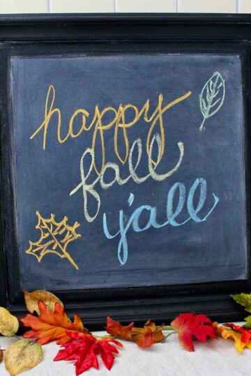 Black framed DIY chalkboard with "happy fall y'all" resting on a counter with fall leaves near by as decoration.