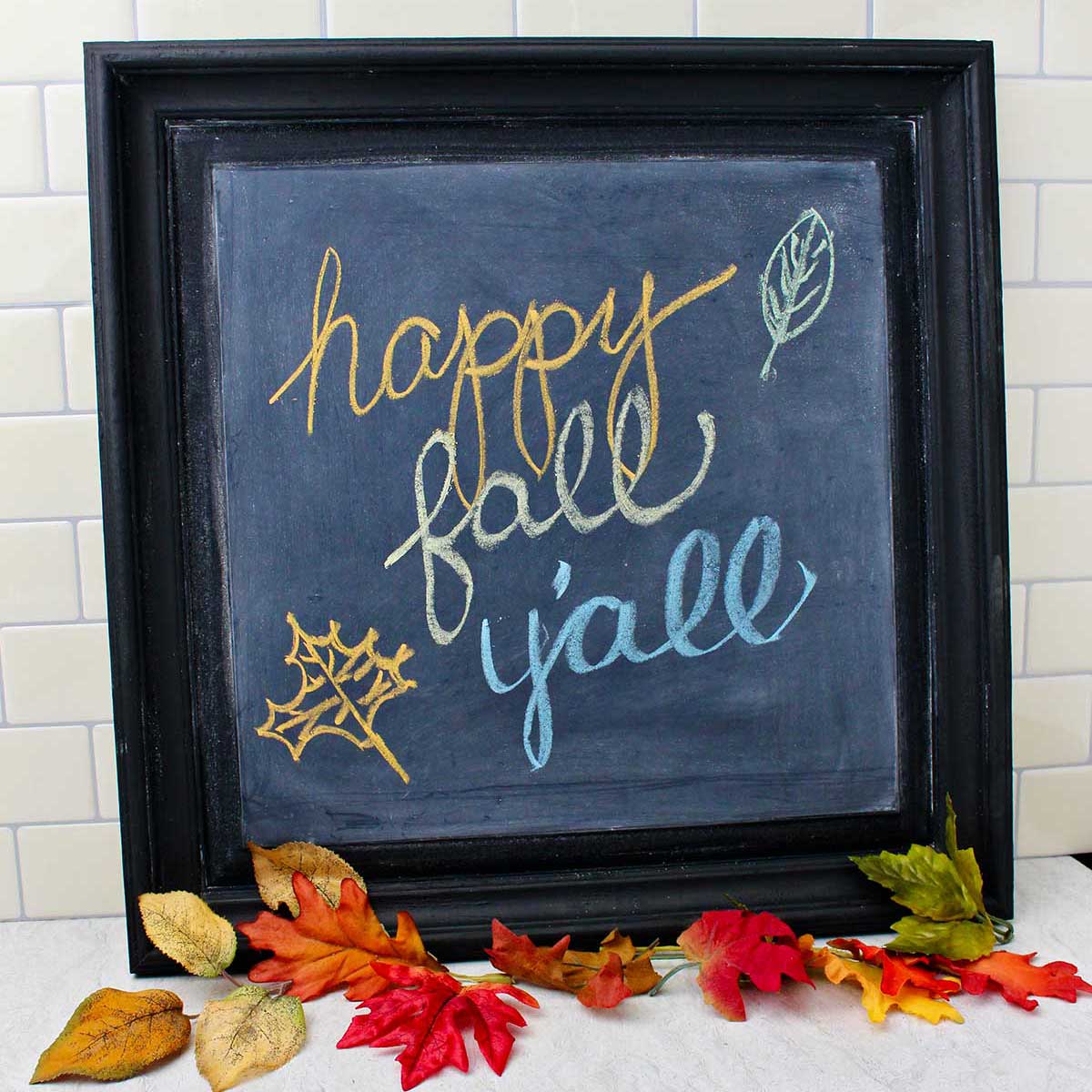 Black framed DIY chalkboard with "happy fall y'all" resting on a counter with fall leaves near by as decoration.