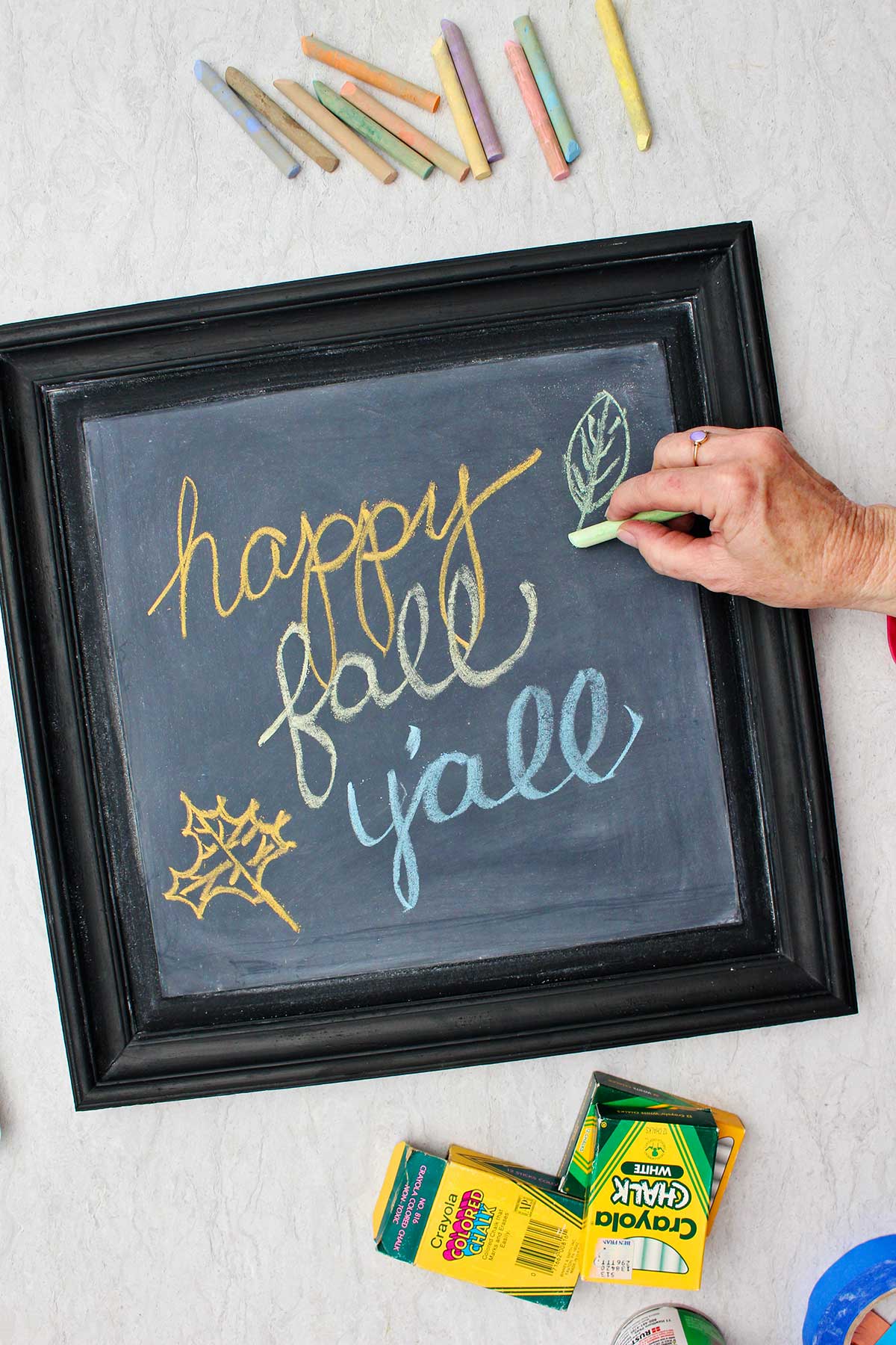 Person writing "happy fall y'all" on DIY chalkboard with boxes of chalk and chalk pieces near by.