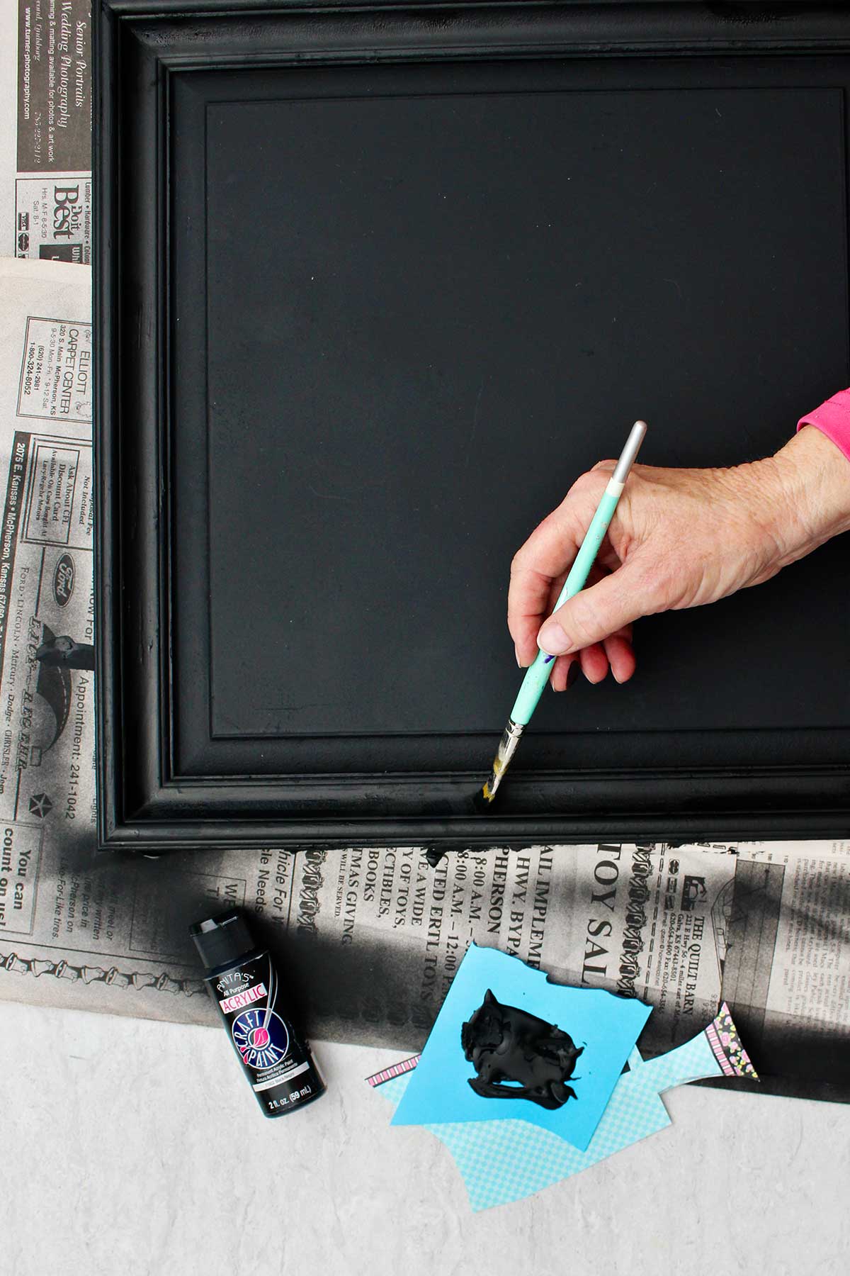 Person painting the frame of DIY chalkboard sign with black acrylic paint.