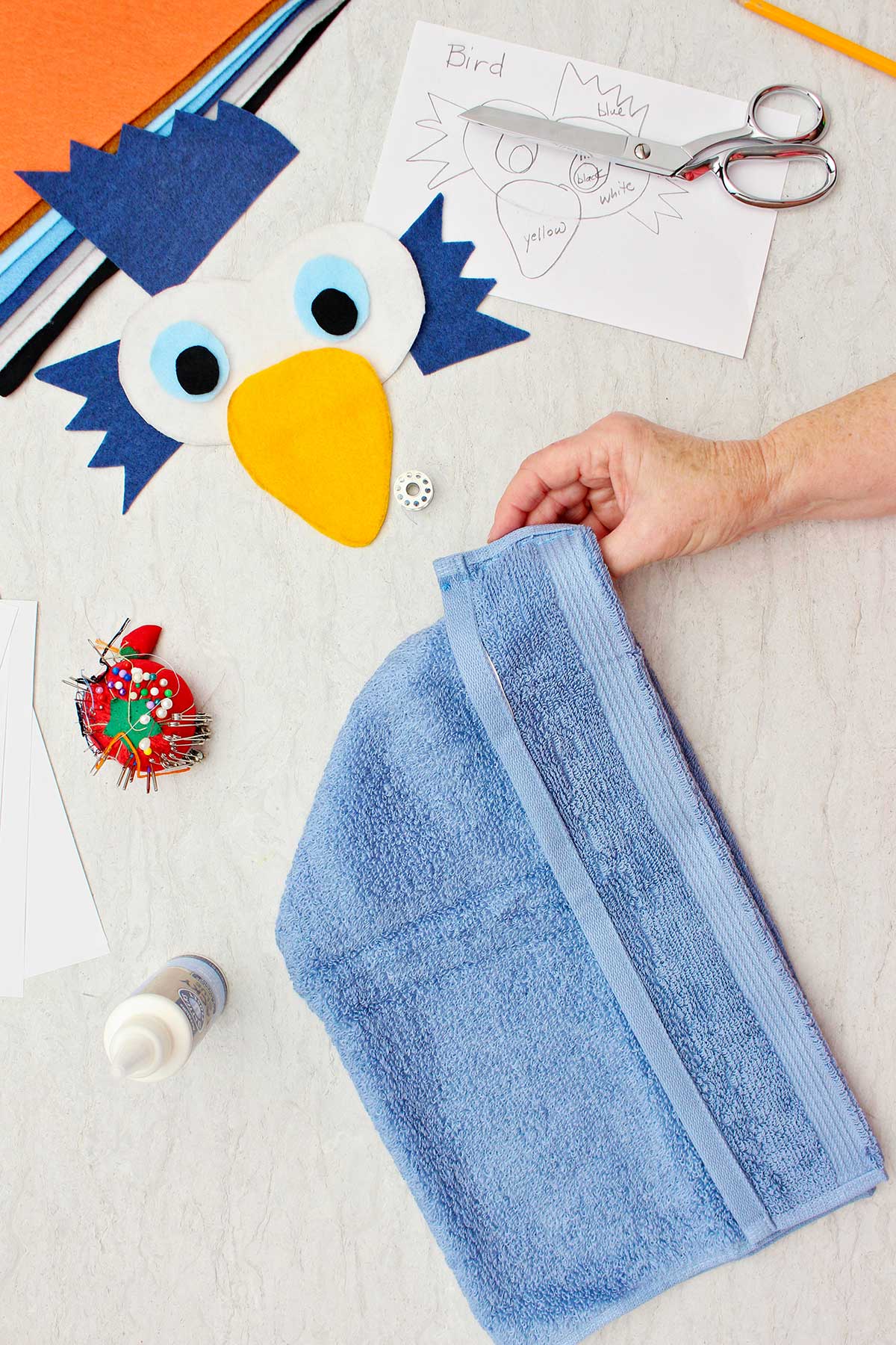 Person holding blue terrycloth towel to make bird costume with some pieces of the bird face cut out near by.