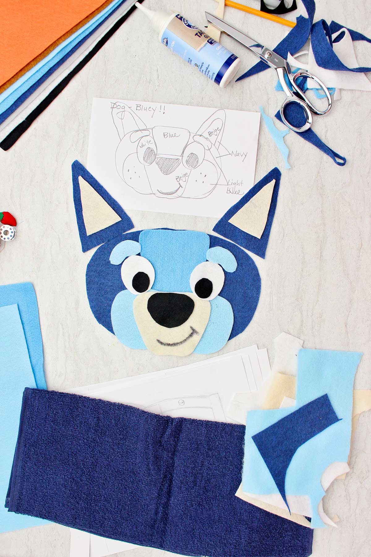 A sketch for a Bluey costume near some cut out pieces of felt for the face and other scraps of supplies near by.