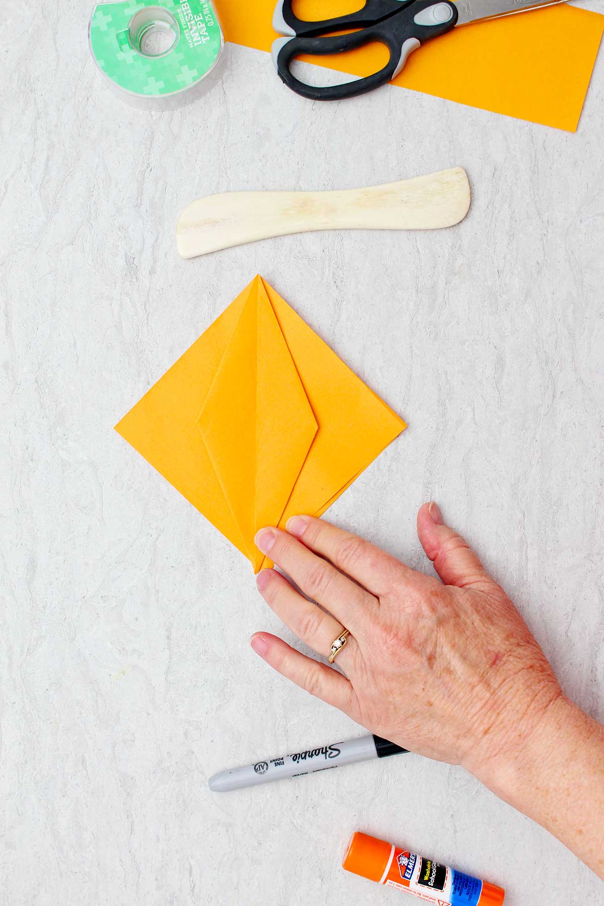 A square piece of orange paper with a folded diamond in the center with other supplies near by to create origami pumpkins.