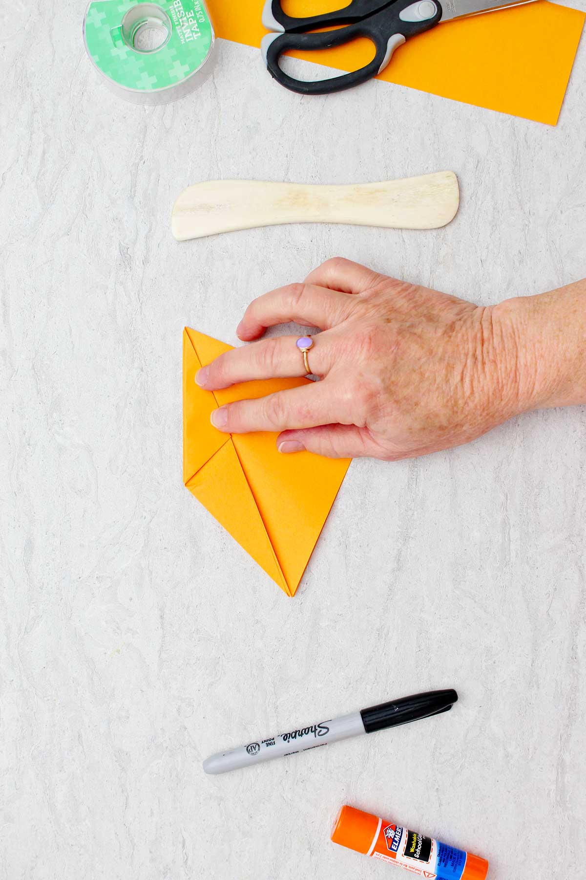 Person folding orange piece of paper to create an origami pumpkin with other supplies near by.