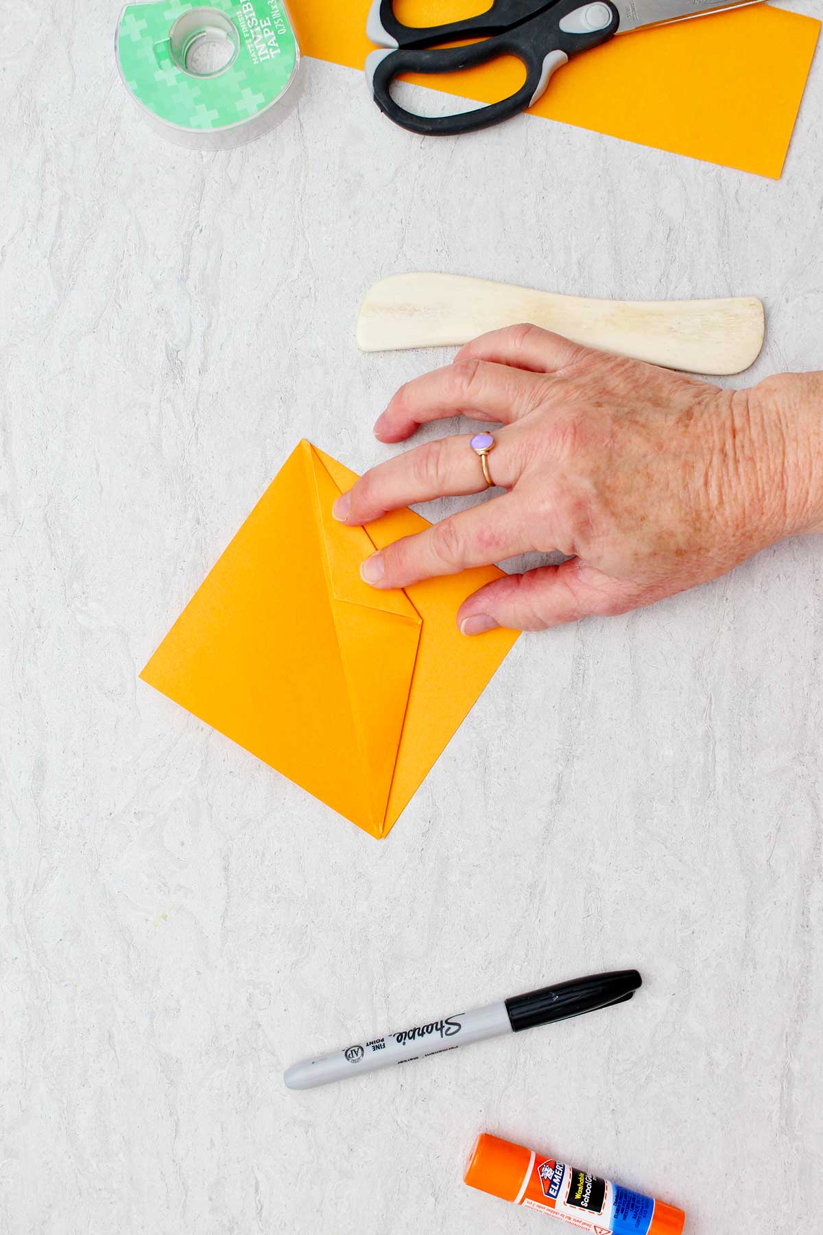 Person folding orange piece of paper to create an origami pumpkin with other supplies near by.