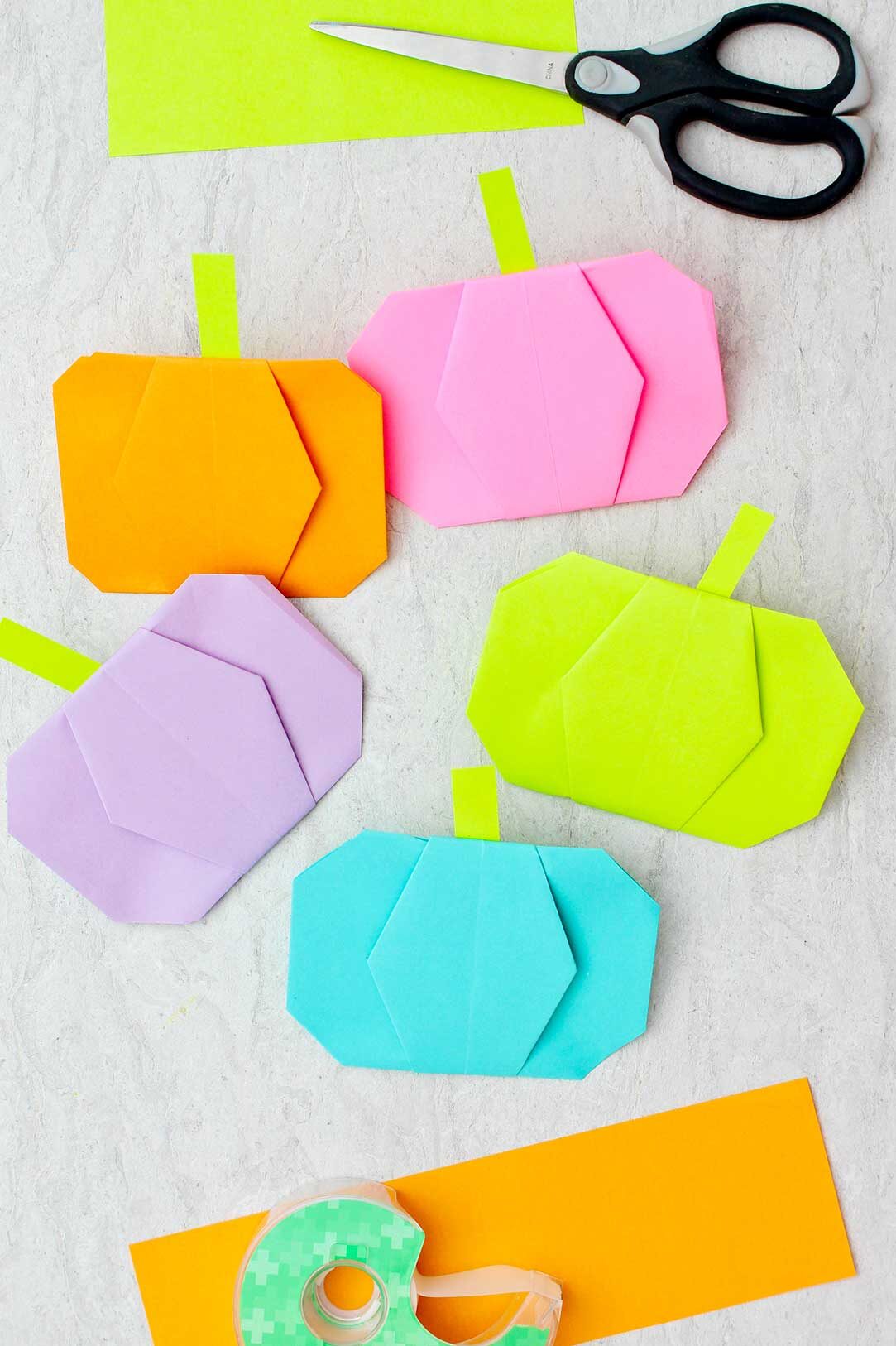 Five different colored origami pumpkins with scissors, tape and other pieces of colorful paper near by.