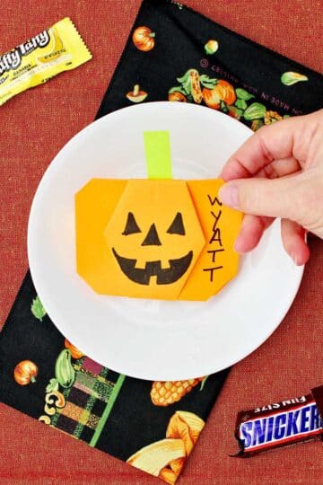 Hand placing an orange origami pumpkin on a place for a place setting with some halloween candy near by.