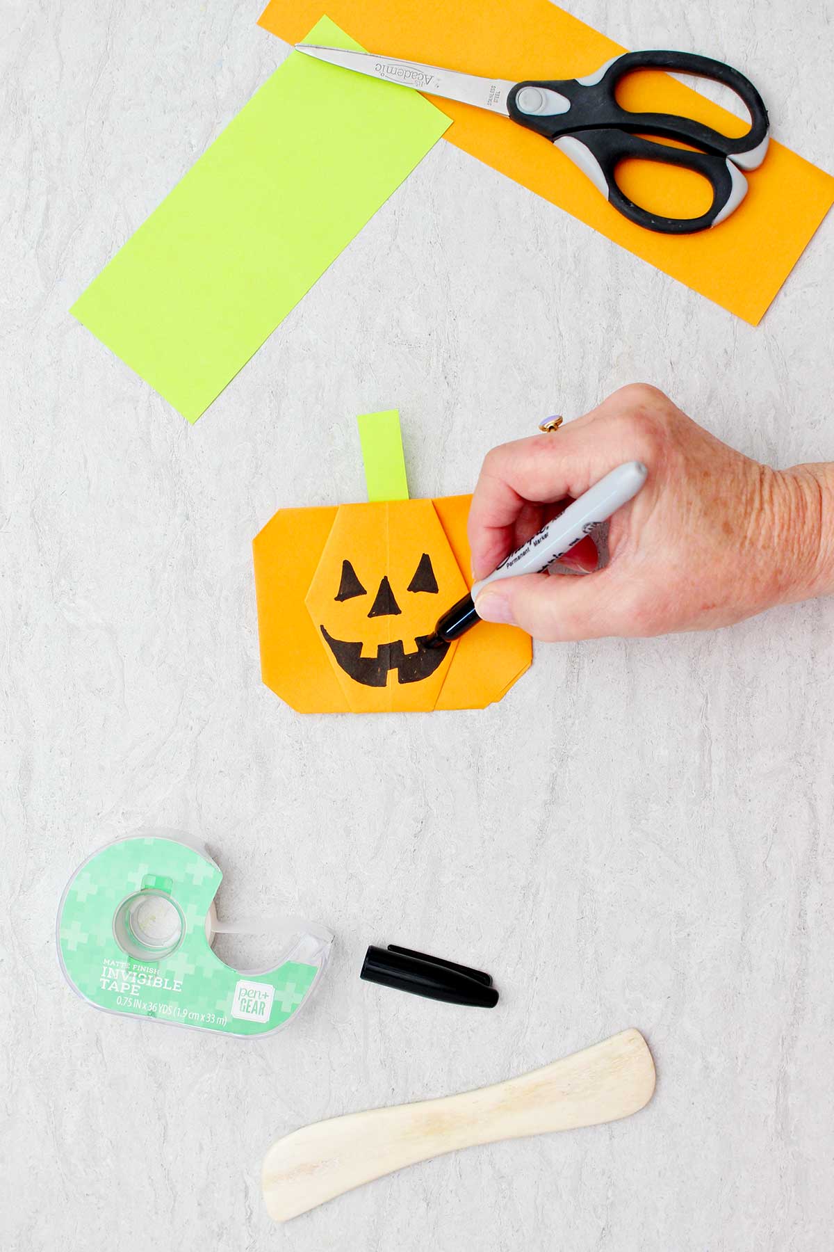 A person drawing a jack-o-lantern face on an orange origami pumpkin with other supplies near by.