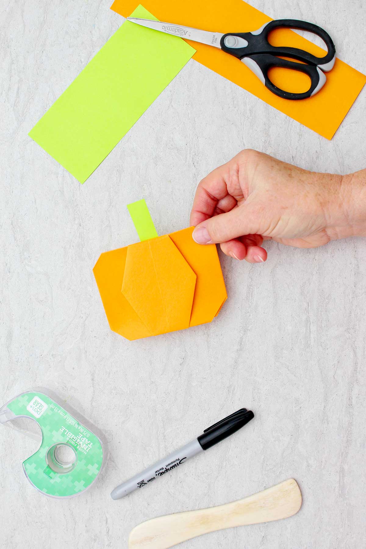 Person holding a completed orange origami pumpkin with a lime green stem with other supplies for making origami pumpkins near by.