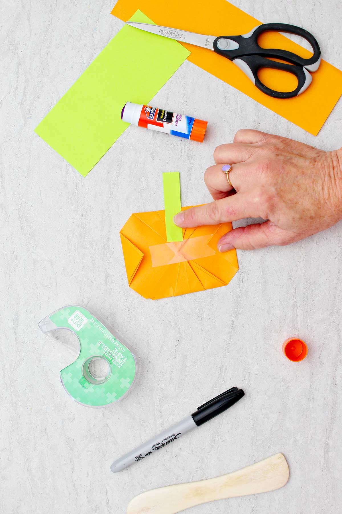 A person taping a green rectangle on the back of an orange origami pumpkin for a stem with other supplies for making origami pumpkins near by.