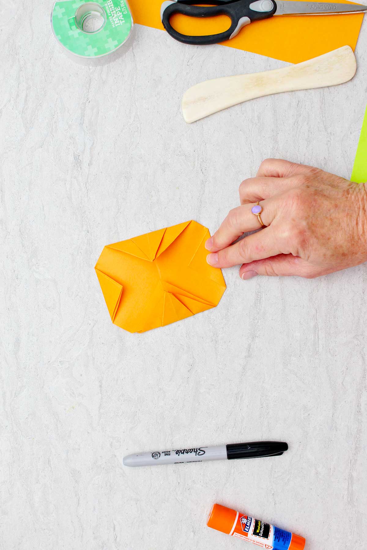 Person folding a corner of orange paper and showing the back side of an almost completed origami pumpkin.