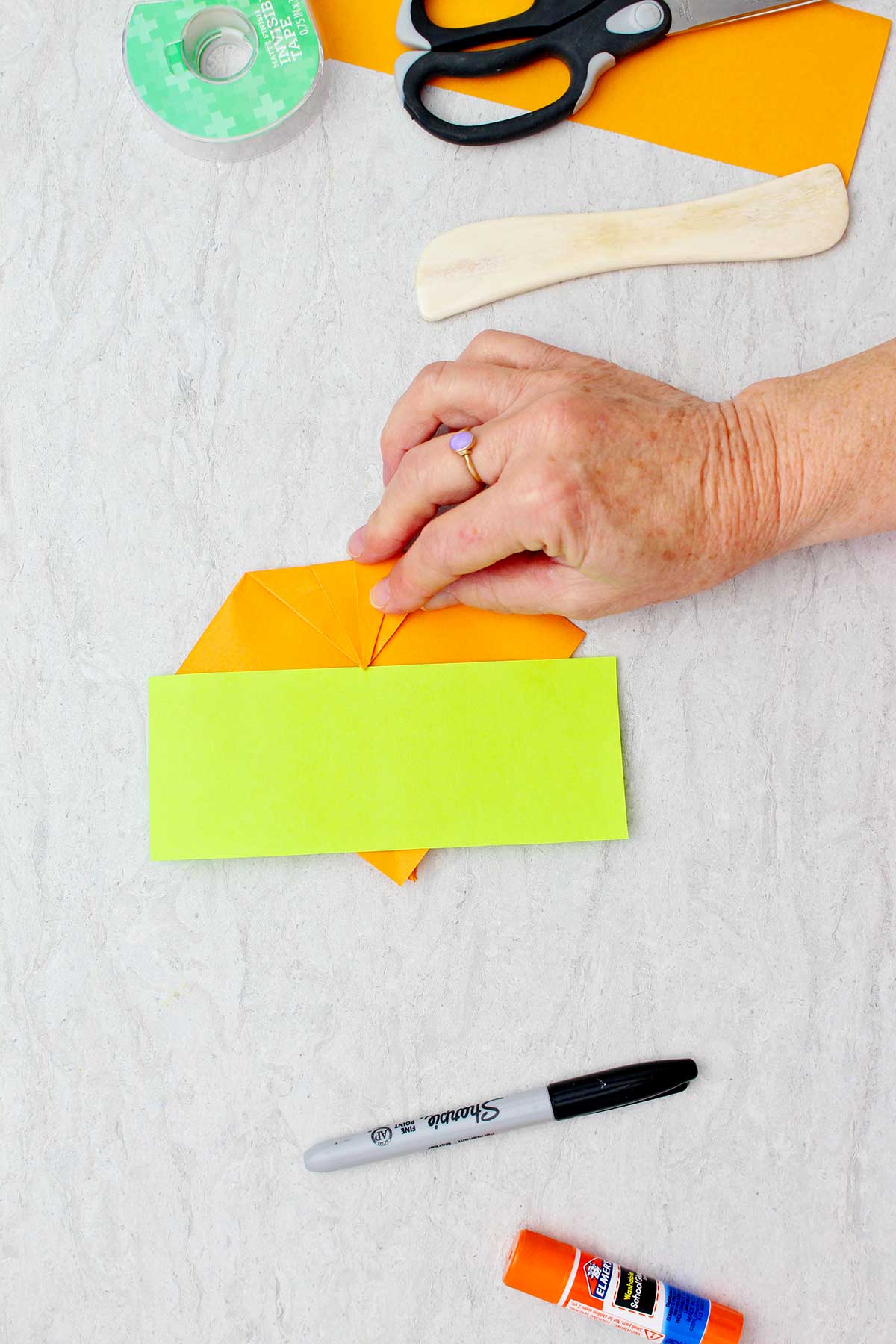 A green rectangle of paper resting on an orange piece of paper that a person is folding with other supplies near by to create origami pumpkins.