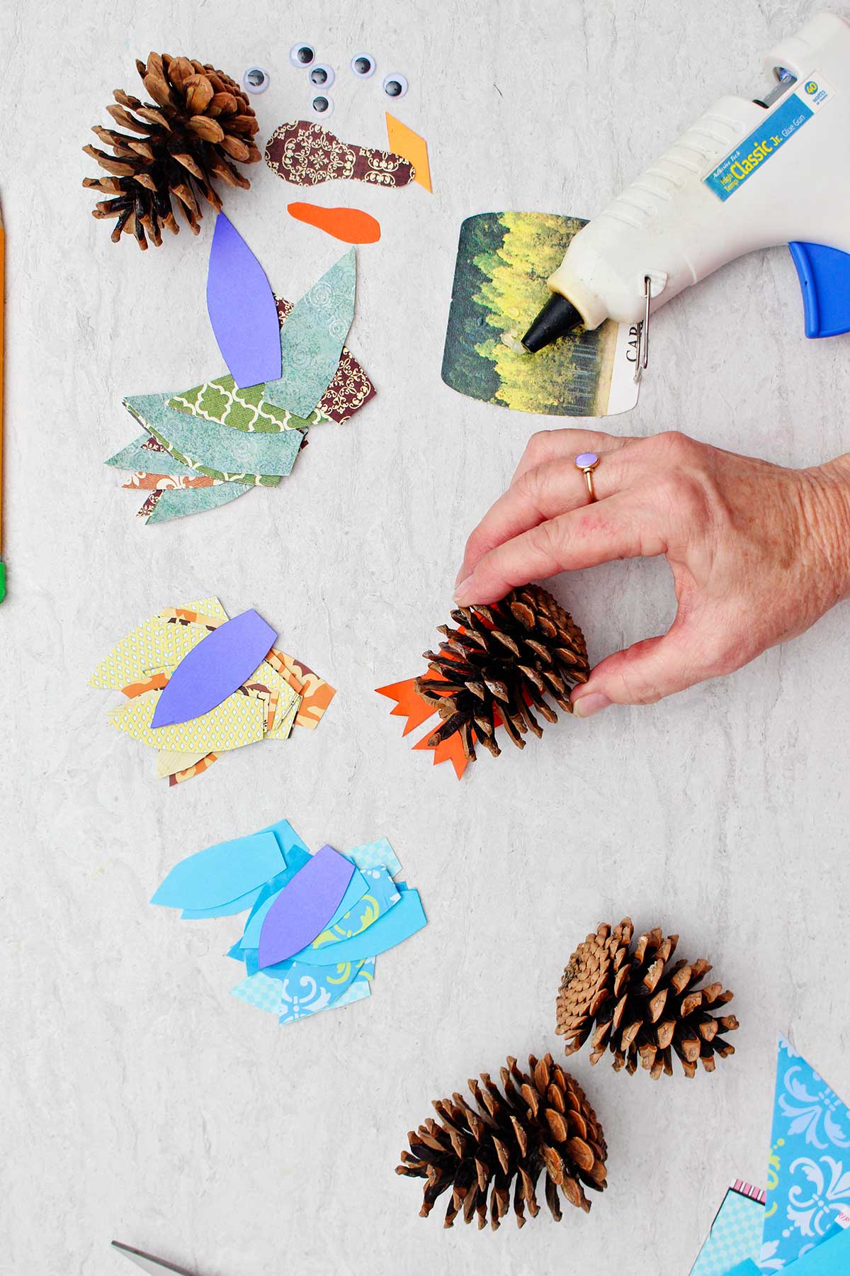 Person gluing a pinecone to paper to make a turkey body with other supplies near by.