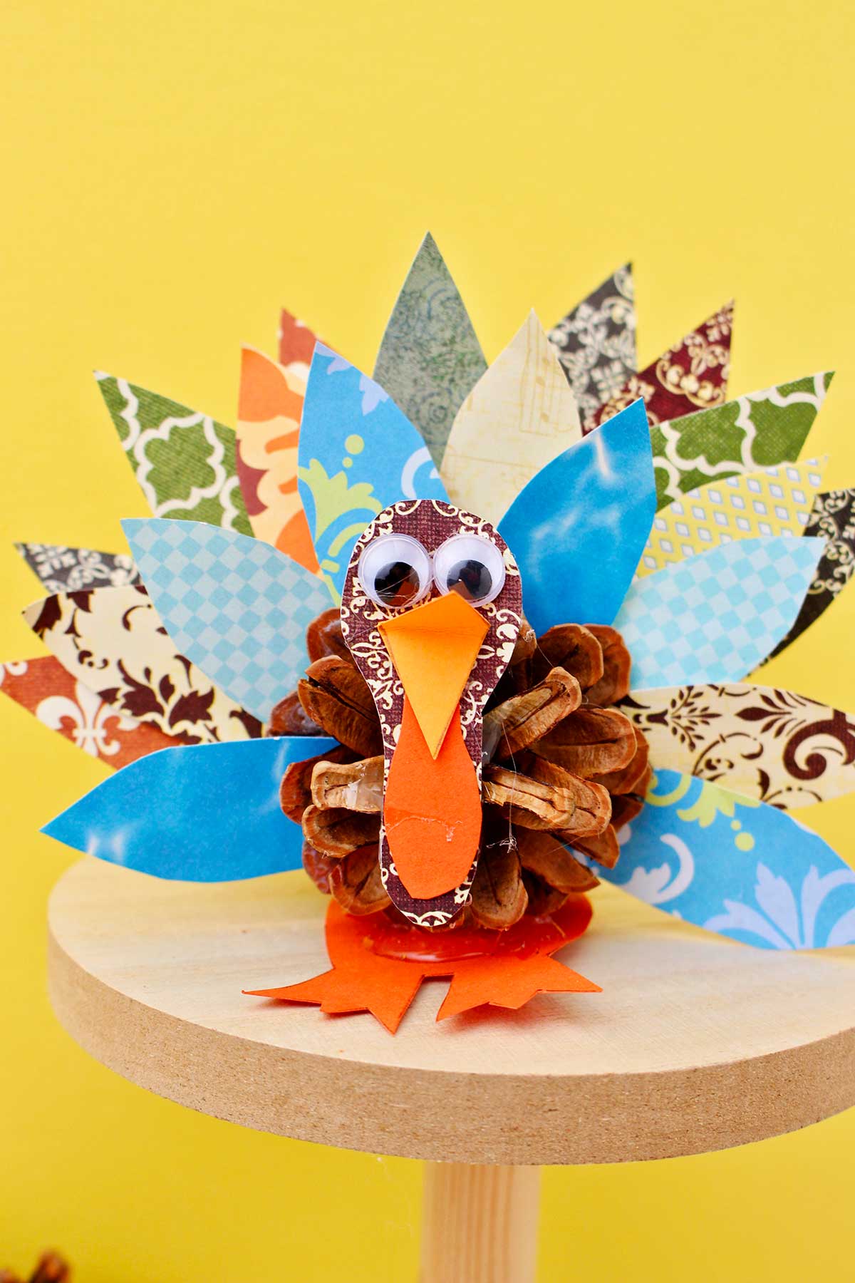 Pinecone turkey made from pattern scrapbook paper, a pinecone and googly eyes on a wooden pedestal against a yellow background.