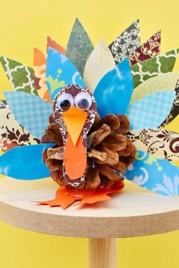 Pinecone turkey made from pattern scrapbook paper, a pinecone and googly eyes on a wooden pedestal against a yellow background.