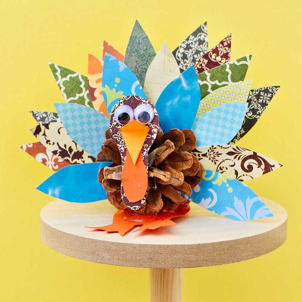 Pinecone turkey made from pattern scrapbook paper, a pinecone and googly eyes on a wooden pedestal against a yellow background.