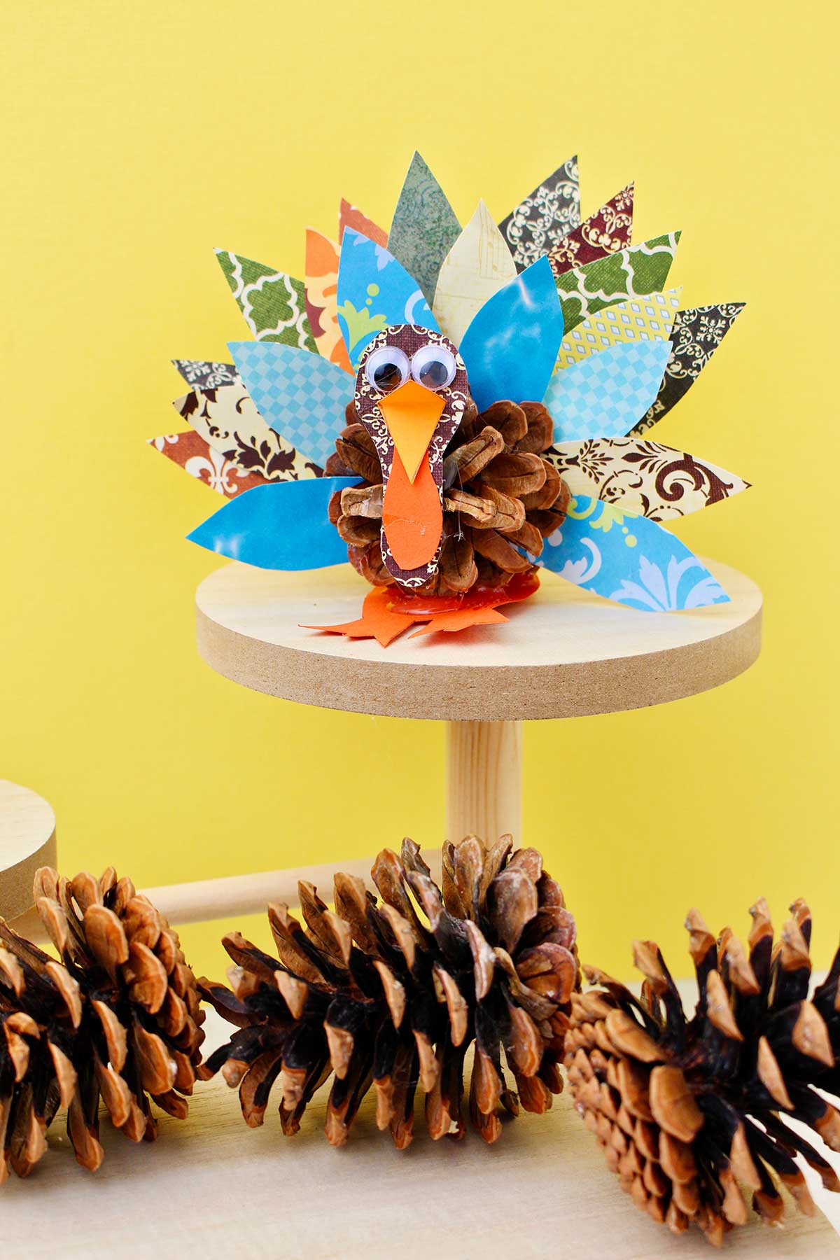 Pinecone turkey made from pattern scrapbook paper, a pinecone and googly eyes on a wooden pedestal against a yellow background with pinecones underneath.