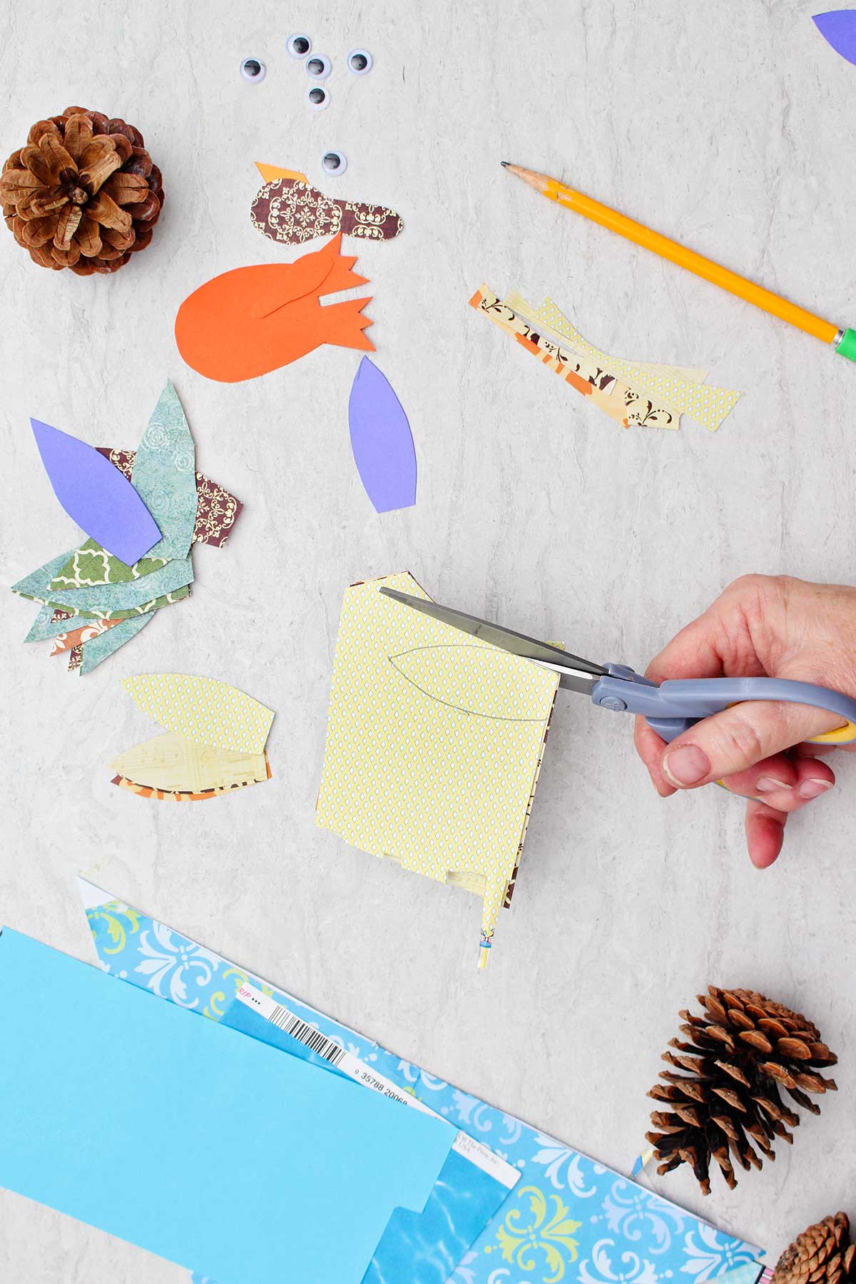 Person cutting a feather out of a pattern yellow piece of scrapbook paper with other supplies near by.