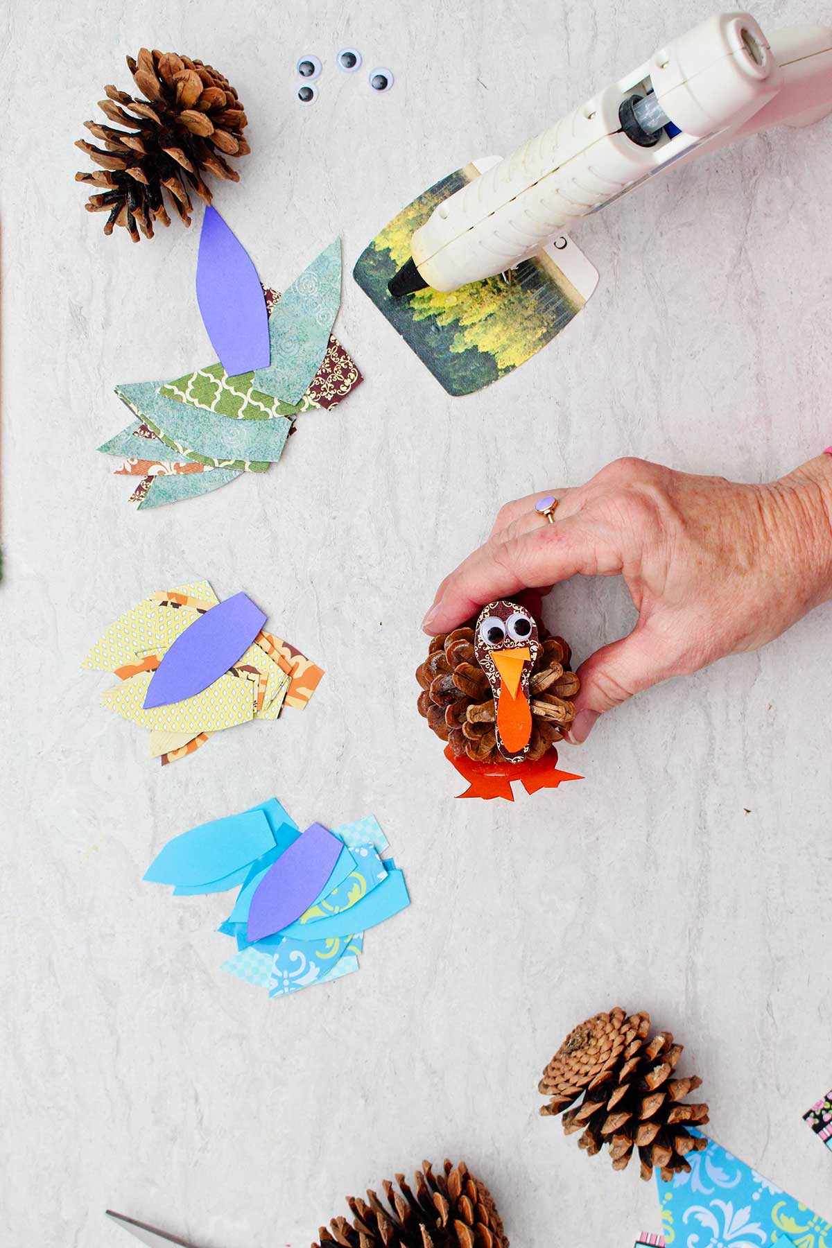 Person holding pinecone with turkey head made from scrapbook paper and google eyes with cut up pieces of scrapbook paper used for feathers near by.