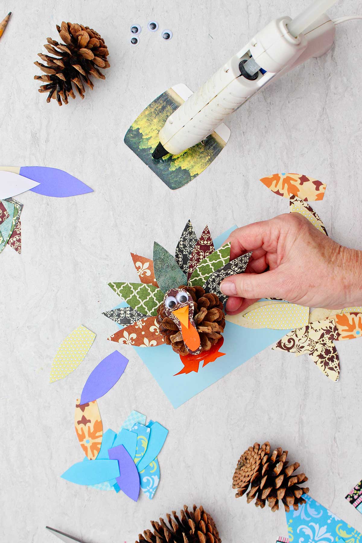 Person gluing a brown pattern scrapbook paper feather on a pinecone turkey with supplies scattered around.