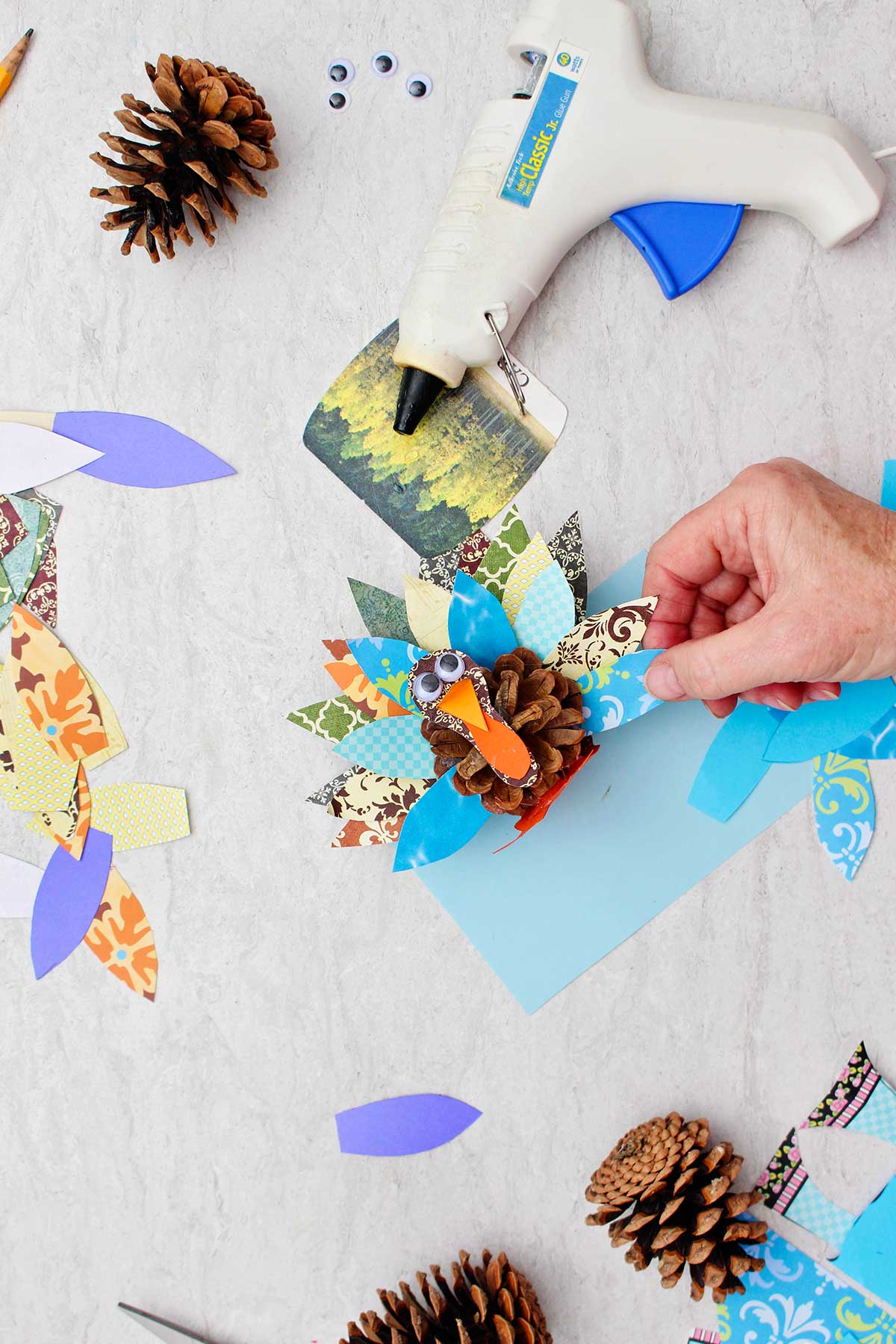 Person gluing a blue scrapbook paper feather on a pinecone turkey with supplies scattered around.