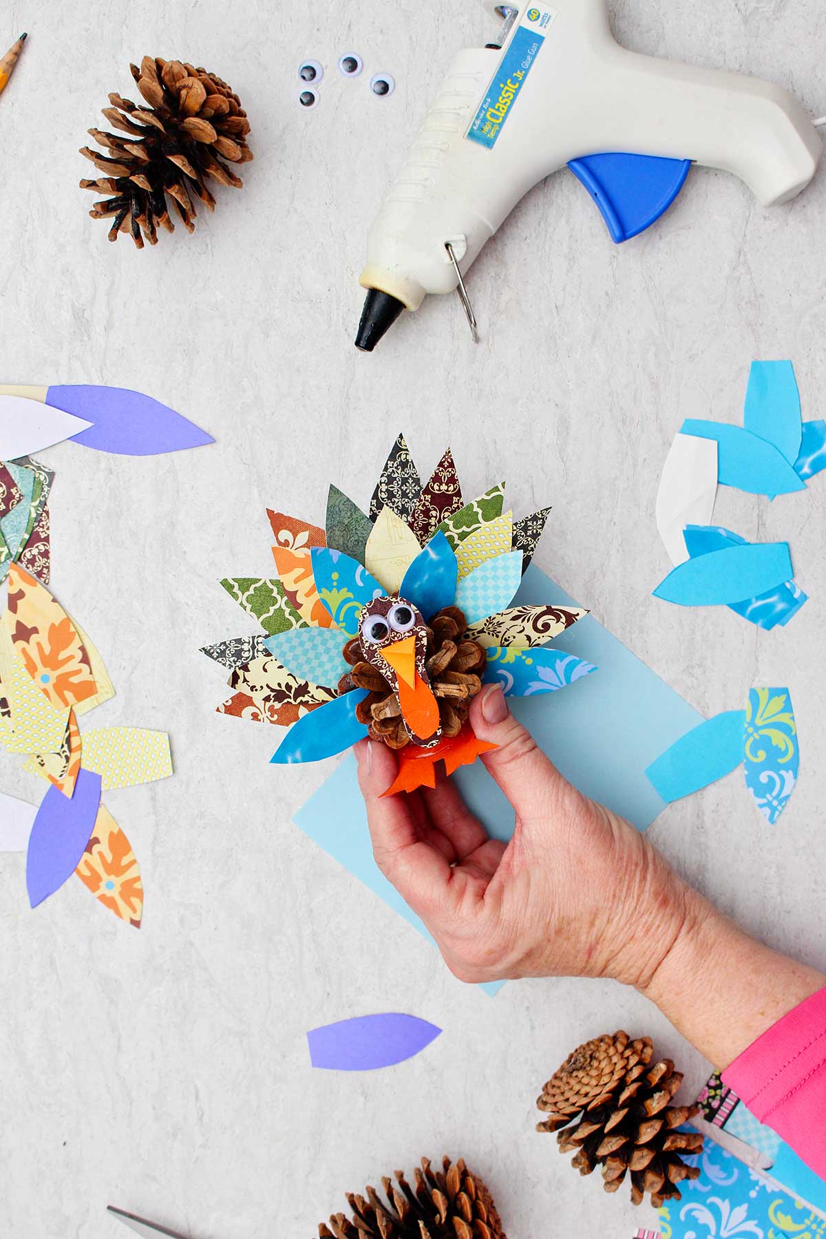 Person holding a completed pinecone turkey made from pattern scrap book paper and a pinecone with pieces of paper, pinecones and a glue gun near by.