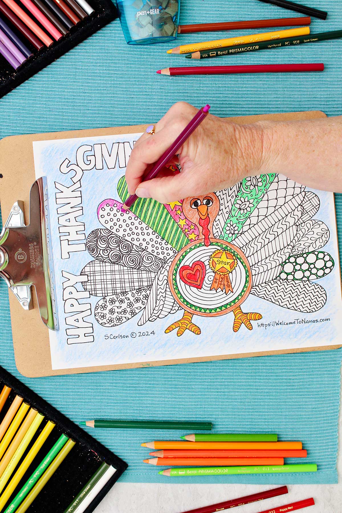 Thanksgiving coloring page of a turkey with patterned wings is clipped into a clipboard and resting on an aqua placemat with colored pencils near by. Person coloring feather magenta.