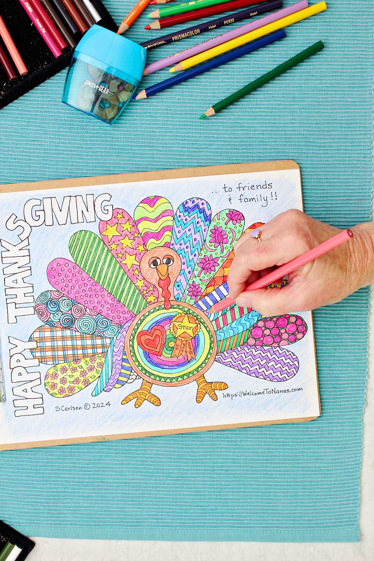 Thanksgiving coloring page of a turkey with patterned wings is clipped into a clipboard and resting on an aqua placemat with colored pencils near by. Person putting finishing touches on turkey feathers.