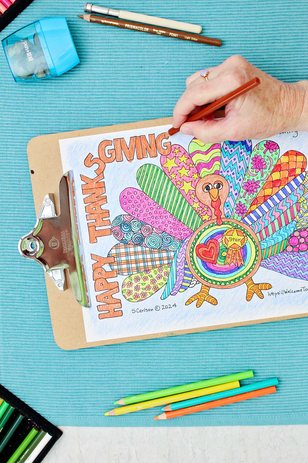 Thanksgiving coloring page of a turkey with patterned wings is clipped into a clipboard and resting on an aqua placemat with colored pencils near by. Person doing some shading in of the turkey's face in brown.