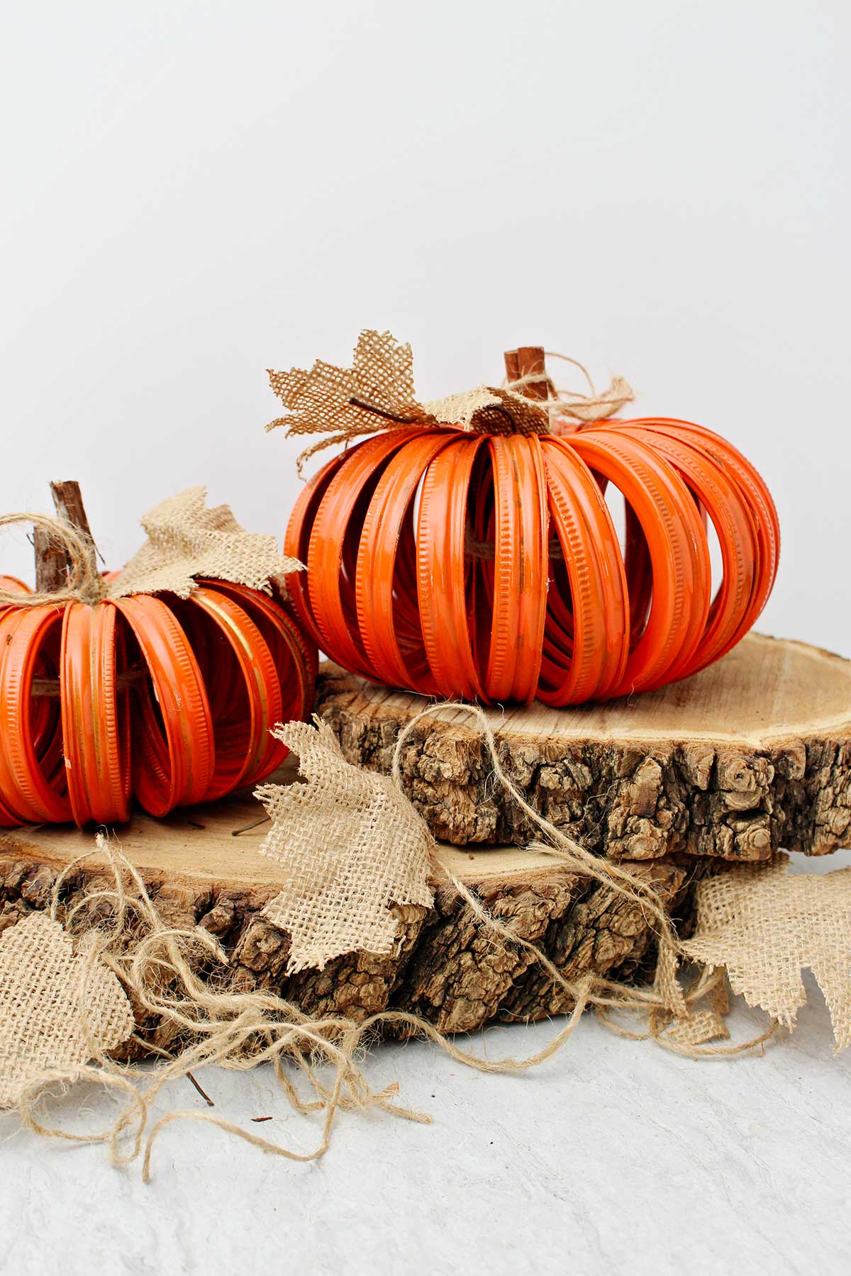 Two completed orange pumpkins made from mason jar lids sitting on two slices of wood with burlap leaves and twine stems.