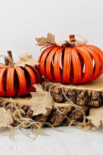 Two completed orange pumpkins made from mason jar lids sitting on two slices of wood with burlap leaves and twine stems.