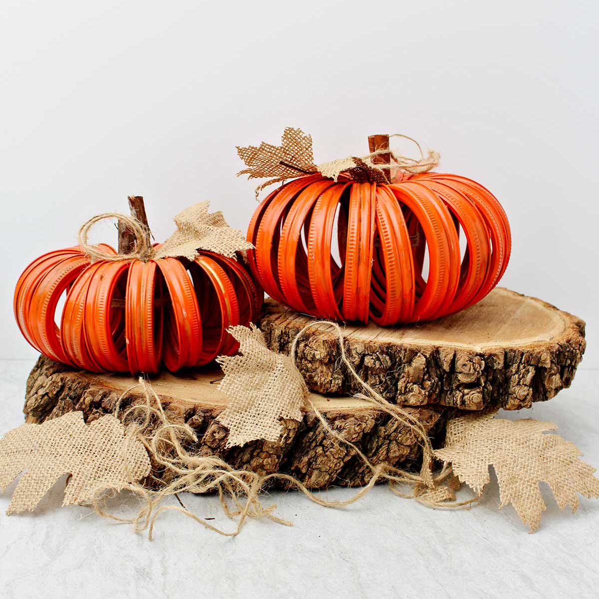 Two completed orange pumpkins made from mason jar lids sitting on two slices of wood with burlap leaves and twine stems.