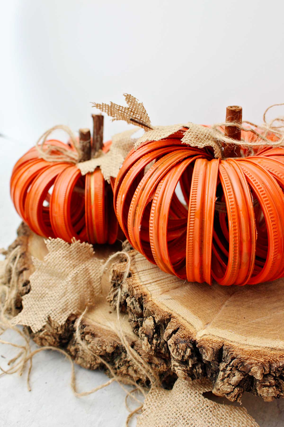 Two completed orange pumpkins made from mason jar lids sitting on two slices of wood with burlap leaves and twine stems.